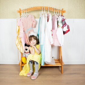 May include: A young girl wearing a yellow dress and a crown sits in front of a wooden clothing rack with a variety of dresses hanging on it. She is holding a stuffed animal and has her eyes closed.