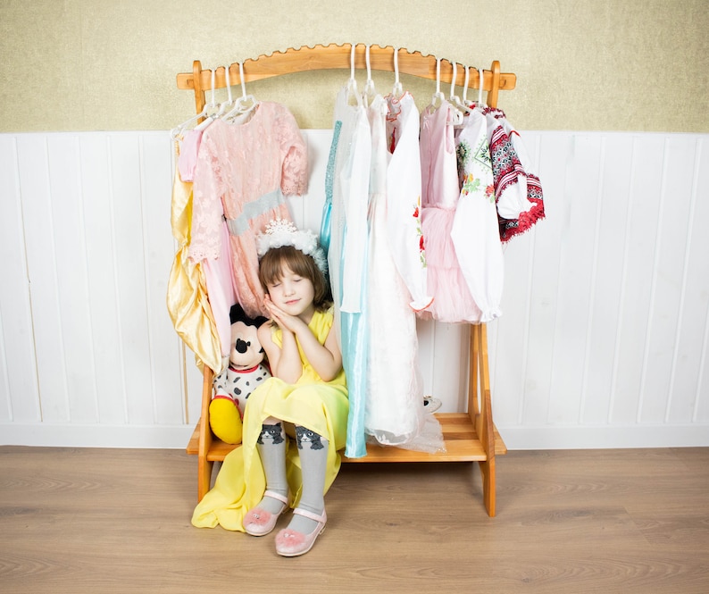 May include: A young girl wearing a yellow dress and a crown sits in front of a wooden clothing rack with a variety of dresses hanging on it. She is holding a stuffed animal and has her eyes closed.