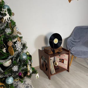 Op de afbeelding: Een bruine houten platenspelerstandaard met een platenspeler erop en platen erin. De standaard heeft een uitgesneden ontwerp aan de zijkant. Een versierde kerstboom is zichtbaar. De standaard is ongeveer 40 cm breed.