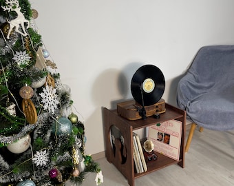 May include: A wooden record player stand with a record player on top and records inside. The stand is brown and has a cutout design on the side. A Christmas tree is decorated with ornaments and lights.