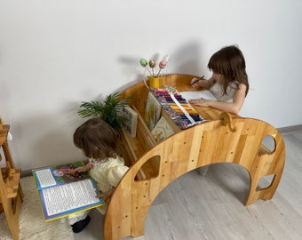 May include: Two children sitting at a wooden table and bookshelf. The table is shaped like a half-circle and has a built-in bookshelf. One child is reading a book and the other is drawing with colored pencils.