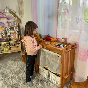 Könnte beinhalten: Eine hölzerne Spielküche mit einem weißen Vorhang, zwei Weidenkörben und einem spielenden Kind. Die Küche hat eine Spüle, ein Kochfeld und Regale. Das Kind trägt ein rosa Hemd und eine schwarze Hose.
