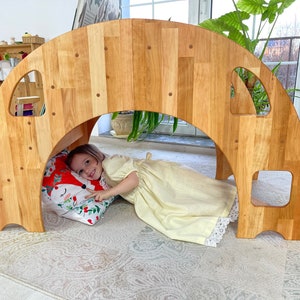 May include: A wooden archway tunnel play structure for children. The archway is made of natural wood and has a rounded top. A child is lying inside the tunnel, looking out.