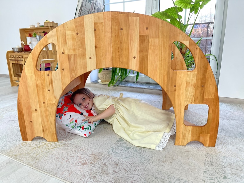 May include: A wooden archway tunnel play structure for children. The archway is made of natural wood and has a rounded top. A child is lying inside the tunnel, looking out.