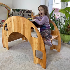 May include: A wooden play structure shaped like a half-circle with a small platform and steps. A young girl is sitting on the platform, coloring in a book.
