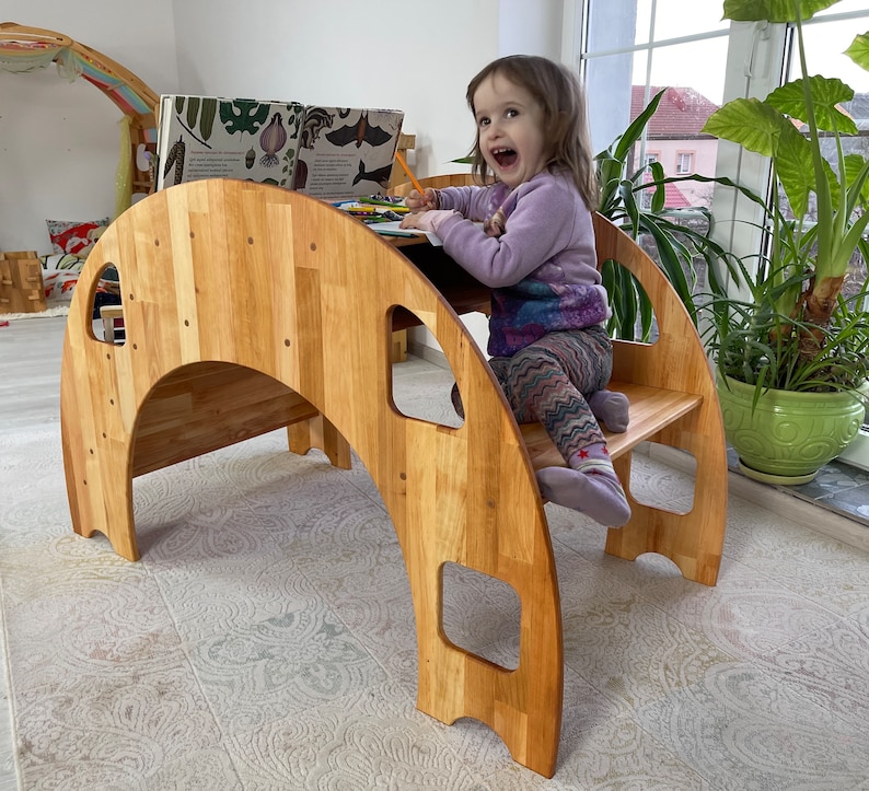 May include: A wooden play structure shaped like a half-circle with a small platform and steps. A young girl is sitting on the platform, coloring in a book.
