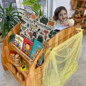 May include: A wooden bookshelf with a yellow fabric canopy, filled with children's books, including "Botanicum" and "Animalum". A young girl sits in front of the bookshelf, reading a book.