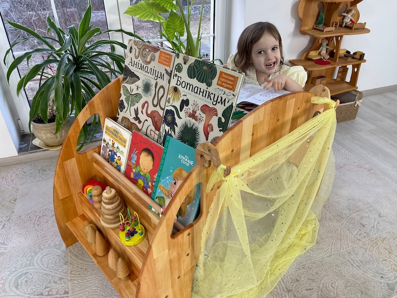 May include: A wooden bookshelf with a yellow fabric canopy, filled with children's books, including "Botanicum" and "Animalum". A young girl sits in front of the bookshelf, reading a book.