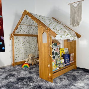 Könnte beinhalten: Holz-Spielhausbett mit Baldachin mit Blättermuster. Das Bett hat ein eingebautes Bücherregal mit Büchern. Ein Teddybär und ein Regenbogenspielzeug befinden sich im Inneren. Das Bett steht auf einem grauen Shaggy-Teppich.