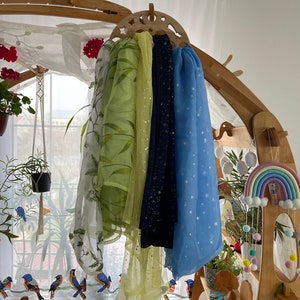 May include: Four sheer scarves hanging on a wooden rack. The scarves are in shades of green, yellow, blue, and black. The scarves have different patterns, including stars, leaves, and glitter.