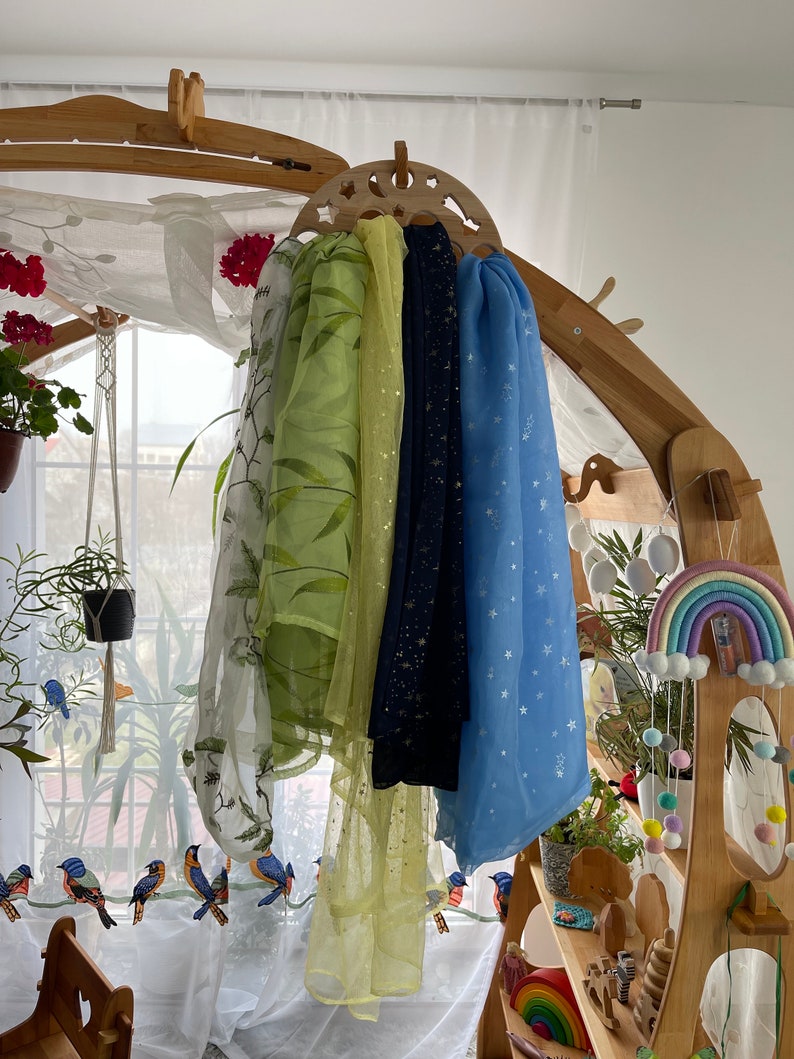 May include: Four sheer scarves hanging on a wooden rack. The scarves are in shades of green, yellow, blue, and black. The scarves have different patterns, including stars, leaves, and glitter.