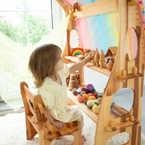 May include: A wooden play kitchen with a rainbow fabric canopy, shelves, and a small wooden chair. A child is playing with wooden toys and fruit.