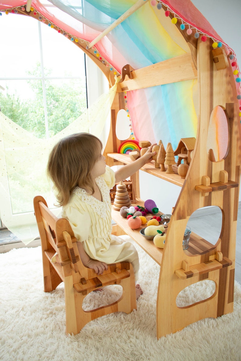 May include: A wooden play kitchen with a rainbow fabric canopy, shelves, and a small wooden chair. A child is playing with wooden toys and fruit.