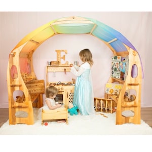 May include: Two children play in a wooden playhouse with a rainbow canopy. The playhouse features a play kitchen, a bookshelf, and a small bed. The playhouse is made of natural wood and has a whimsical design.