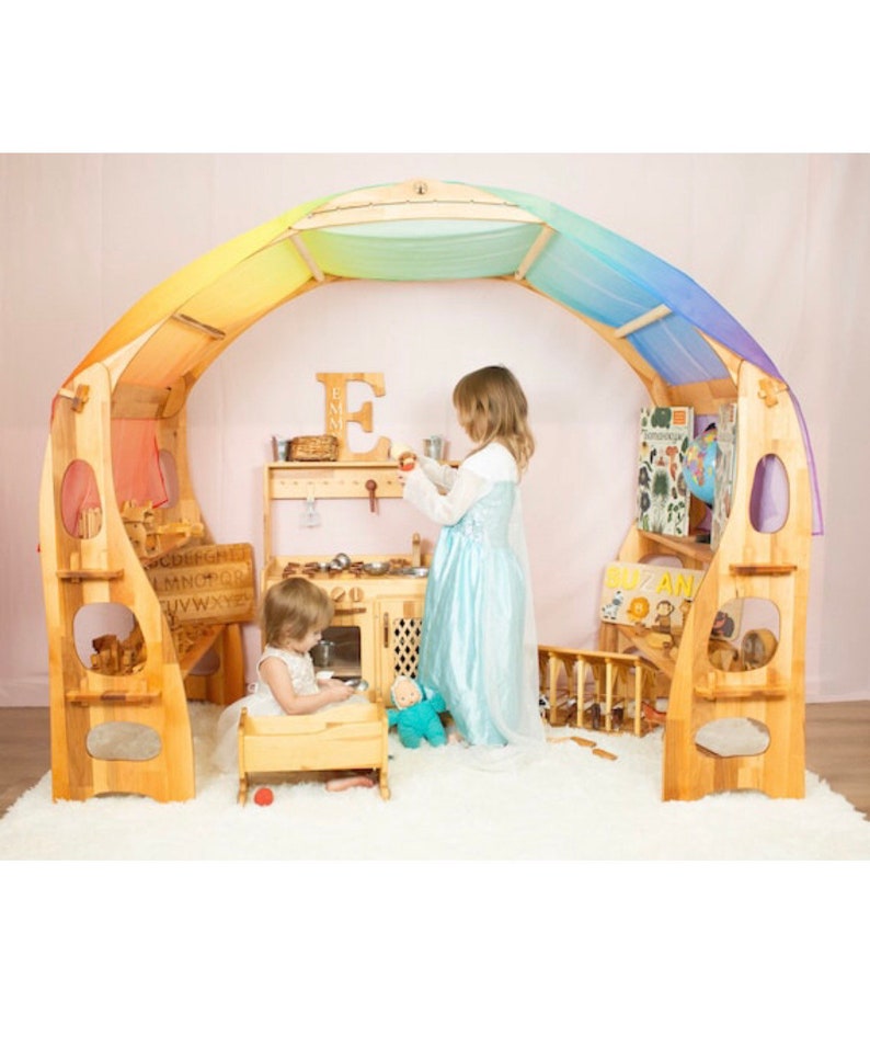 May include: Two children play in a wooden playhouse with a rainbow canopy. The playhouse features a play kitchen, a bookshelf, and a small bed. The playhouse is made of natural wood and has a whimsical design.