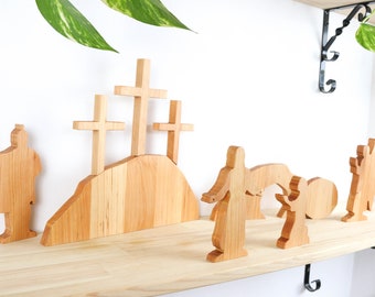 May include: Wooden Easter scene featuring three crosses on a hill, figures of people, and an empty tomb. The figures are made of light-colored wood and are arranged on a wooden shelf. The scene is a religious themed decoration.