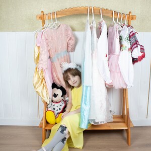 May include: A young girl sits in front of a wooden clothes rack with a variety of dresses hanging on it. She is wearing a yellow dress and grey tights with a cat design. A stuffed Mickey Mouse toy sits next to her.