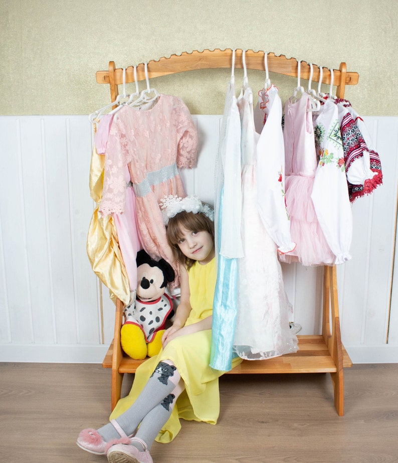 May include: A young girl sits in front of a wooden clothes rack with a variety of dresses hanging on it. She is wearing a yellow dress and grey tights with a cat design. A stuffed Mickey Mouse toy sits next to her.