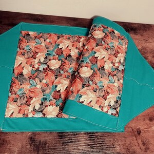 Op de afbeelding: Een turquoise tafelkleed met een gestikt ontwerp en een herfstbladerenprint in tinten oranje, bruin en crème.