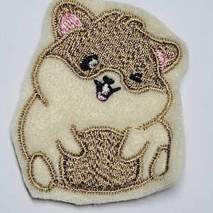 May include: Embroidered patch featuring a cartoon hamster design. The hamster has brown fur, pink ears, and black eyes, outlined with gold thread. The patch is on a cream-colored felt background.