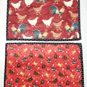 May include: Two red quilted placemats with a chicken and egg design. The top placemat features a vintage style print with chickens and roosters. The bottom placemat has a repeating pattern of chickens and eggs.