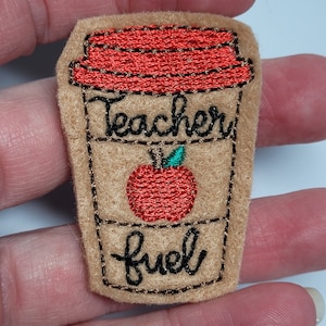 May include: A brown felt patch with embroidered text that reads "Teacher fuel" and a red apple. The patch is shaped like a coffee cup with a red lid.