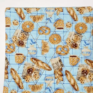 May include: A blue quilted hot pad with a pattern of brown bread, pastries, coffee mugs, and cookies. The pattern is repeated on a light blue background with a grid pattern.