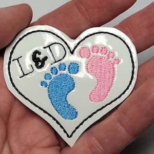 May include: White heart-shaped patch with black stitching around the edges. The patch features the letters "L & D" in black embroidery and two embroidered footprints, one blue and one pink.