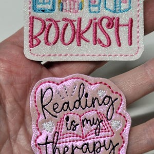 May include: Two embroidered patches with book-themed designs. One patch is rectangular with the word "BOOKISH" in pink, and icons of a tablet, headphones, a coffee cup, and an open book. The other patch is pink and reads "Reading is my therapy."