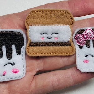 May include: Three felt appliques shaped like s'mores. Each applique has a white marshmallow with a brown chocolate topping and a smiling face. The middle applique has a brown graham cracker on top and bottom. The applique on the right has a pink bow on top.