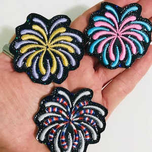 May include: Three embroidered iron-on patches in the shape of fireworks. The first patch is purple and gold, the second is pink and blue, and the third is red, white, and blue.