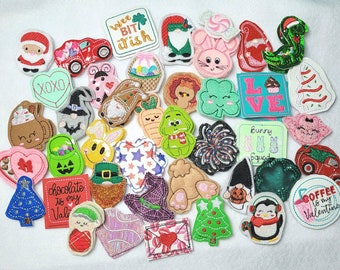 May include: A collection of felt appliques featuring various designs, including hearts, gnomes, a Christmas tree, a penguin, a cupcake, a bunny, a shamrock, a pumpkin, a candy cane, a firecracker, a star, a gingerbread man, a carrot, a witch, a reindeer, a truck, a dinosaur, a snowman, a present, a coffee cup, and a candy heart. The appliques are all brightly colored and have a whimsical feel.
