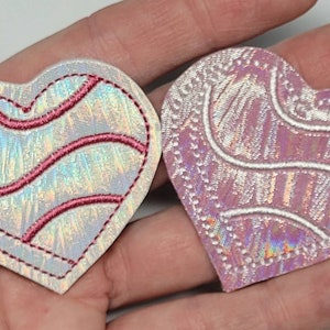 May include: Two iridescent heart-shaped patches with pink and white embroidery. The patches have a white outline and a pink swirl design.