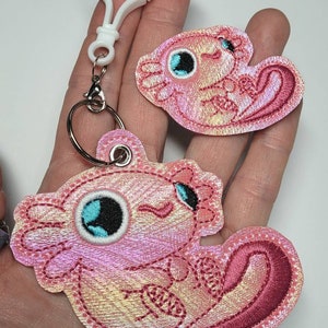 May include: A pink and white axolotl keychain with iridescent fabric. The axolotl is embroidered with a cute expression and has a white ring around its eye. The keychain has a white plastic clip and a silver metal ring.