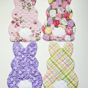 May include: Four quilted Easter bunny shapes in different fabric patterns. The bunnies are pink with floral patterns, pink with a tulip pattern, purple with a floral pattern, and pink and green with a plaid pattern.