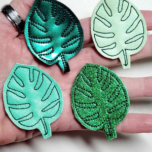 May include: Four green monstera leaf fabric patches. Two are a light green with a dark green outline. Two are a dark green with a light green outline. One of the dark green patches has a glittery texture.
