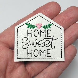 May include: White felt house-shaped patch with black embroidery that reads "Home Sweet Home" with a pink flower and green leaves above the text.