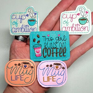 May include: Five embroidered iron-on patches with coffee cup designs. The patches are in various colors, including pink, orange, white, and turquoise. The patches feature text such as "cup of ambition", "This girl runs on coffee", and "Mug Life".