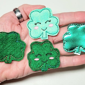 May include: Four felt shamrock patches, two green, one teal, and one green with a shiny finish. Two of the green shamrocks have a smiling face with pink cheeks.