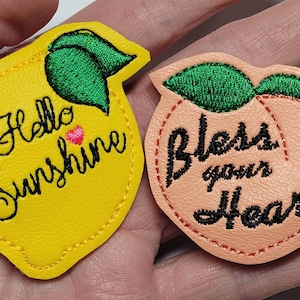 May include: Two embroidered patches: a yellow lemon with "Hello Sunshine" and a peach with "Bless your Heart". The lemon has green leaves and a pink heart. The peach has green leaves and a red stitched outline.