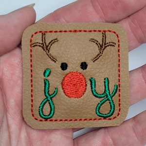 May include: A brown leather patch with red stitching around the edges. The patch features an embroidered reindeer with a red nose and the word "joy" in green.