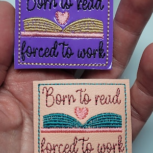 May include: Two embroidered patches with the text "Born to read, forced to work." One patch is purple with gold and black stitching, the other is peach with teal and red stitching. Both feature an open book and heart.