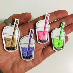 May include: Embroidered patches in the shape of bubble tea cups, each with a different colored drink and straw. The drinks are brown, purple, pink, and green. The patches have a white background and a black border with a stitched design.
