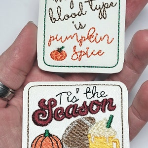 May include: Two embroidered patches with fall themes. The first patch is white with green stitching and says "My blood type is pumpkin spice" with a small orange pumpkin. The second patch is white with brown stitching and says "Tis' the season" with a football, a pumpkin, and a cup of hot chocolate.
