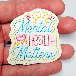 May include: White embroidered patch with a yellow outline. The patch features a sun, a heart, and the text "Mental Health Matters" in blue, pink, and yellow thread.