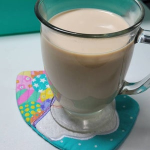 May include: A glass mug filled with a light brown beverage, sitting on a colorful quilted coaster with a white cloud design.