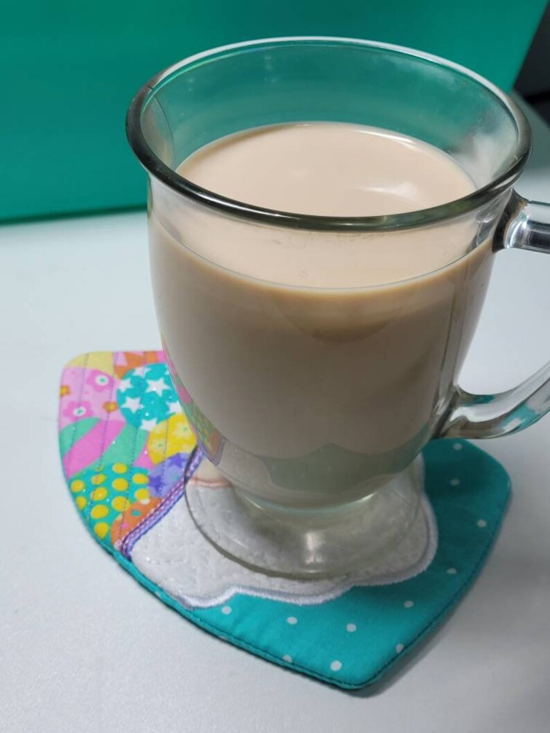 May include: A glass mug filled with a light brown beverage, sitting on a colorful quilted coaster with a white cloud design.