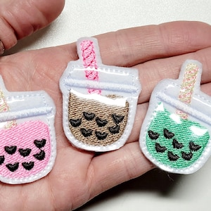 May include: Three embroidered iron-on patches of bubble tea cups with different colored drinks. The patches are white with a clear plastic cup design, a pink drink with black boba, a brown drink with brown boba, and a green drink with black boba. Each patch has a pink and white striped straw.