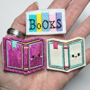 May include: Three embroidered book-themed patches. One rectangular patch with the word "BOOKS" and colorful vertical stripes. Two open book patches, one pink with glitter and one white, each with a bookmark and a smiling face.