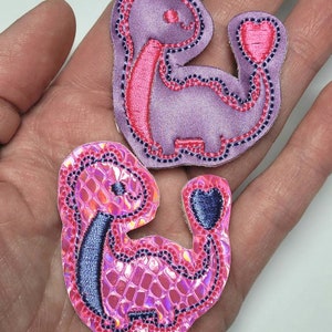 May include: Two embroidered iron-on patches featuring pink and purple dinosaurs holding hearts. The dinosaur on the left has a pink iridescent finish and the dinosaur on the right is a solid purple.